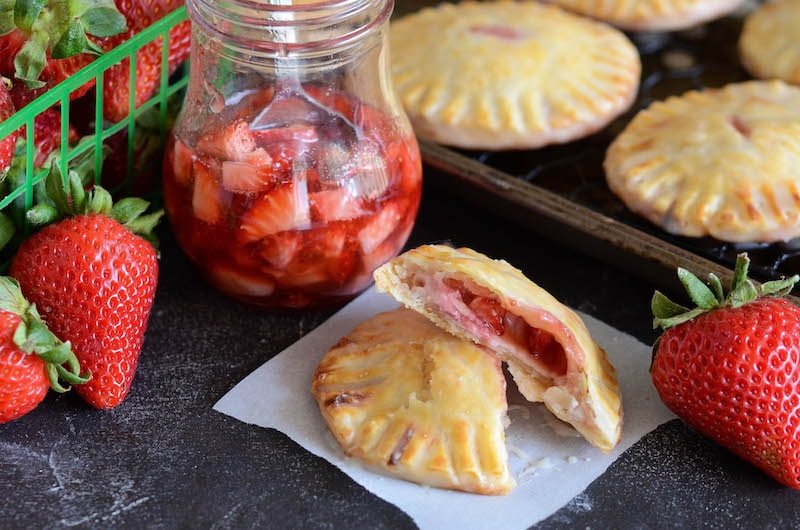 Two halves of a strawberry and cream hand pie on a serviette, surrounded by a jar of strawberry filling and fresh strawberries.