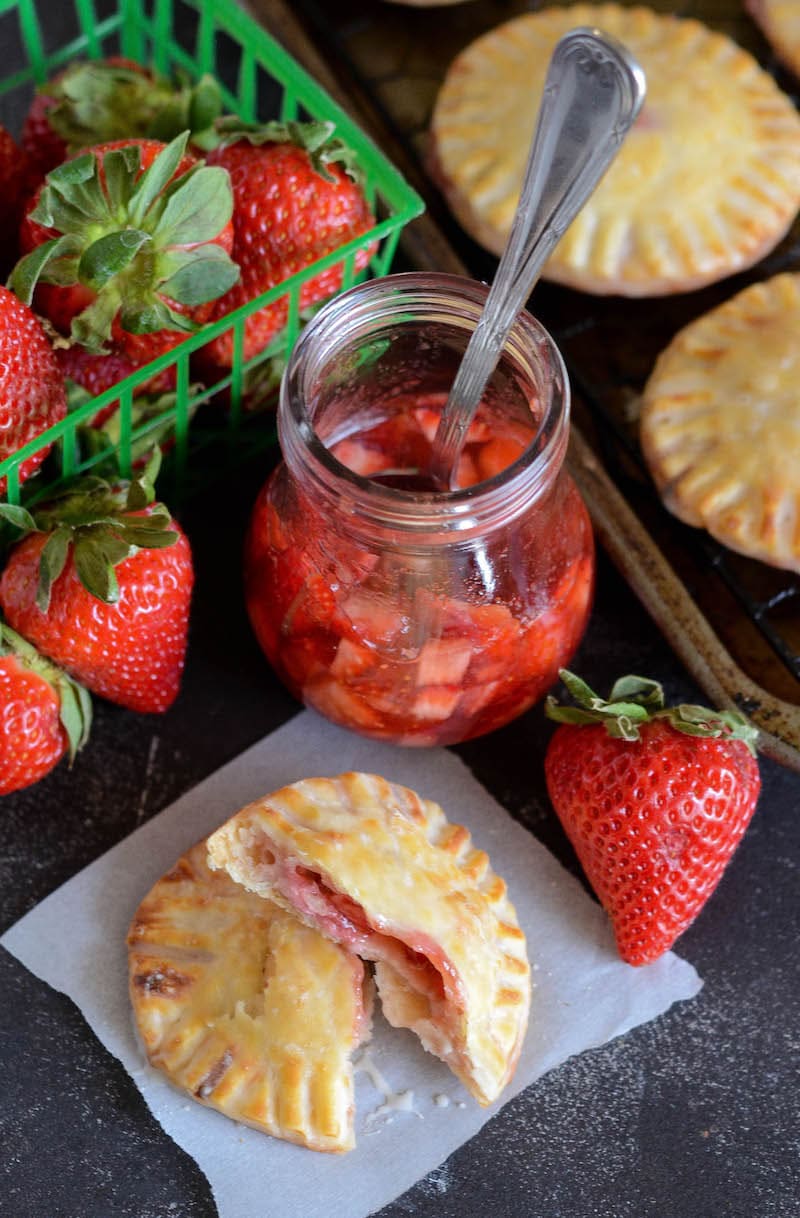 Two halves of a strawberry and cream hand pie on a serviette, surrounded by a jar of strawberry filling and fresh strawberries.