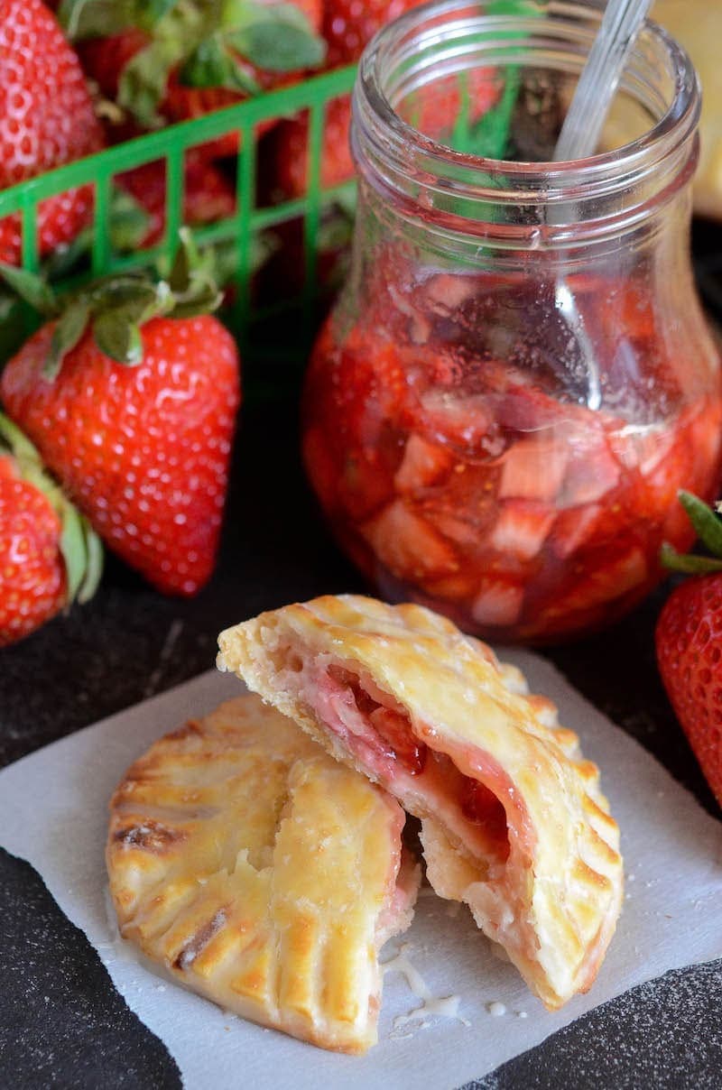 Two halves of a strawberry and cream hand pie on a serviette, surrounded by a jar of strawberry filling and fresh strawberries.
