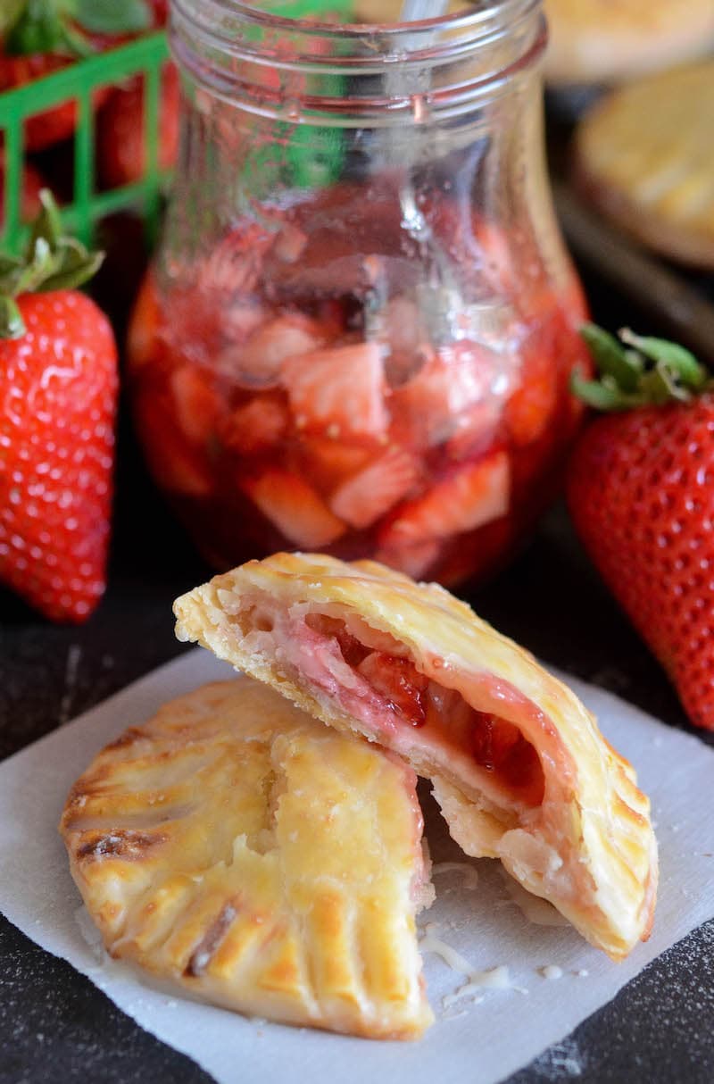 Two halves of a strawberry and cream hand pie on a serviette, surrounded by a jar of strawberry filling and fresh strawberries.