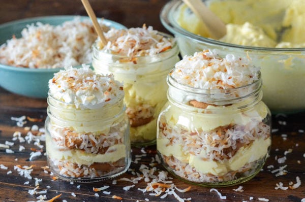 Mason jars with layers of coconut pudding in front of a large mixing bowl and a bowl of toasted coconut.