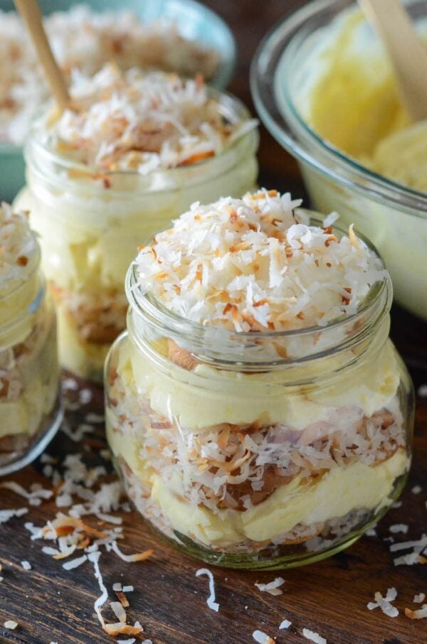 Three mason jars filled with layers of vanilla pudding, Nilla wafers and toasted coconut in front of a large mixing bowl. 