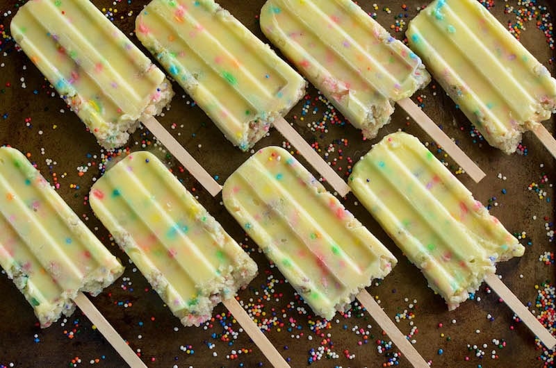 Eight Funfetti Cake Pudding Pops Lined Up on a Baking Sheet with Loose Nonpareil Sprinkles