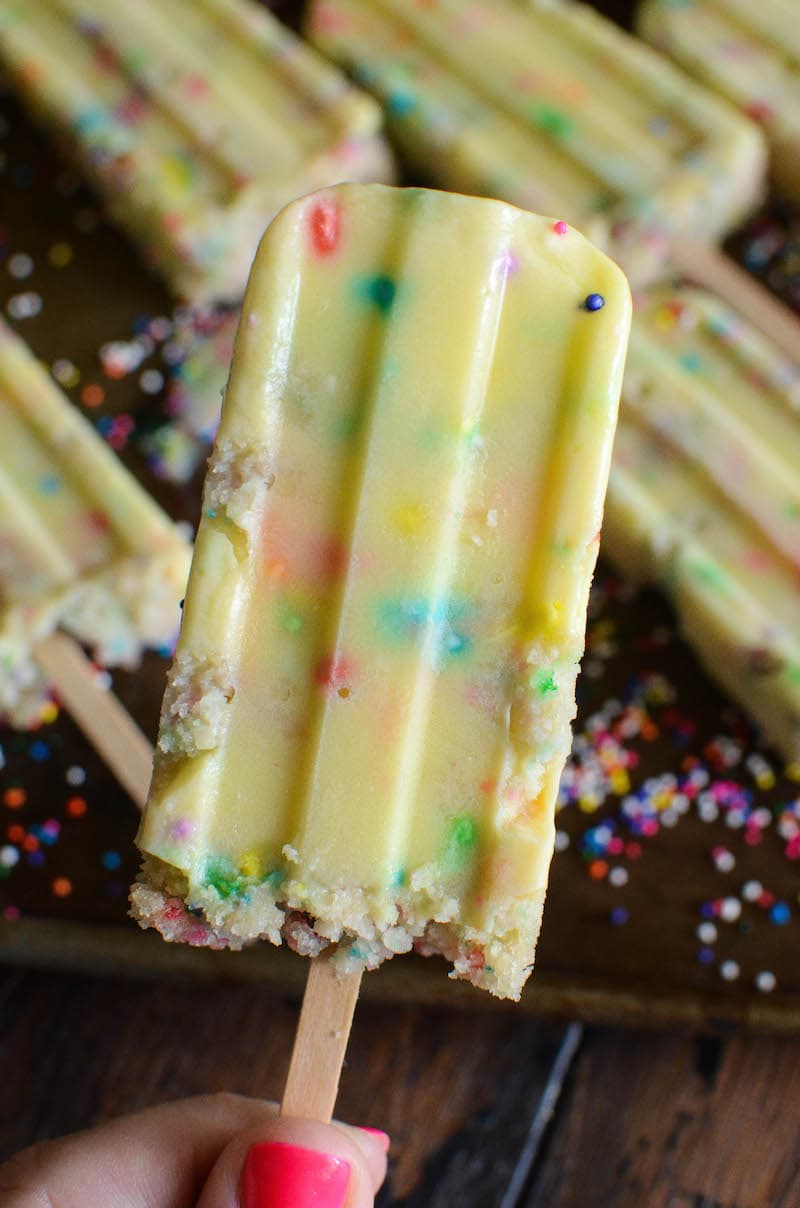 A Funfetti Cake Pudding Pop Being Held Above a Baking Sheet Holding More Popsicles