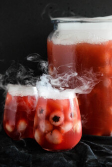 Glasses of sparkling cranberry halloween eyeball punch with floating lychees, next to a punch bowl garnished with dry ice.