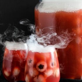 Glasses of sparkling cranberry halloween eyeball punch with floating lychees, next to a punch bowl garnished with dry ice.