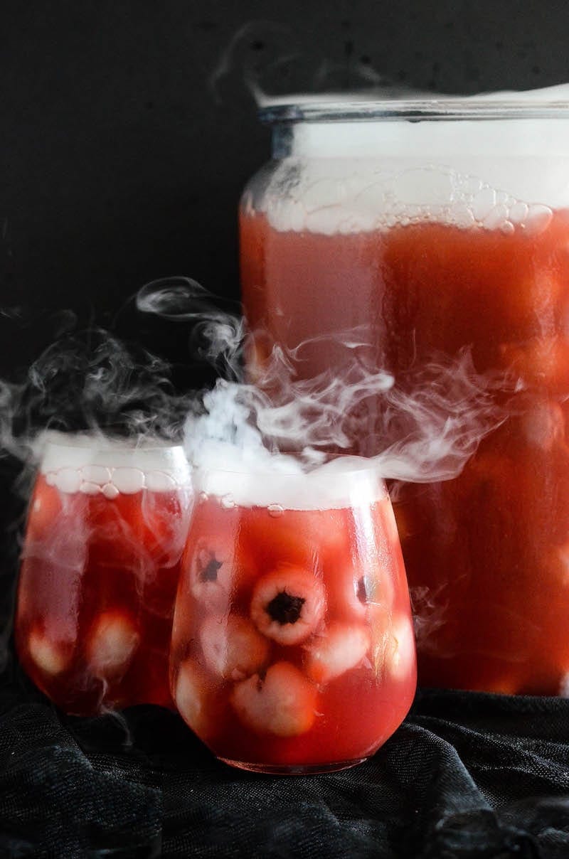 Glasses of sparkling cranberry halloween eyeball punch with floating lychees, next to a punch bowl garnished with dry ice.