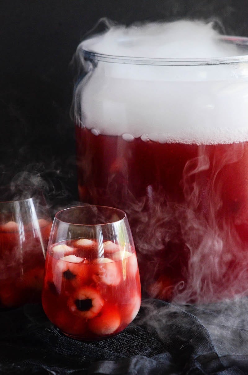 Glasses of sparkling cranberry halloween eyeball punch with floating lychees, next to a punch bowl garnished with dry ice.