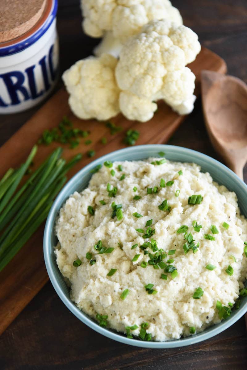 A bowl of prepared cauliflower mash next to the ingredients: cauliflower, chopped chives, and garlic