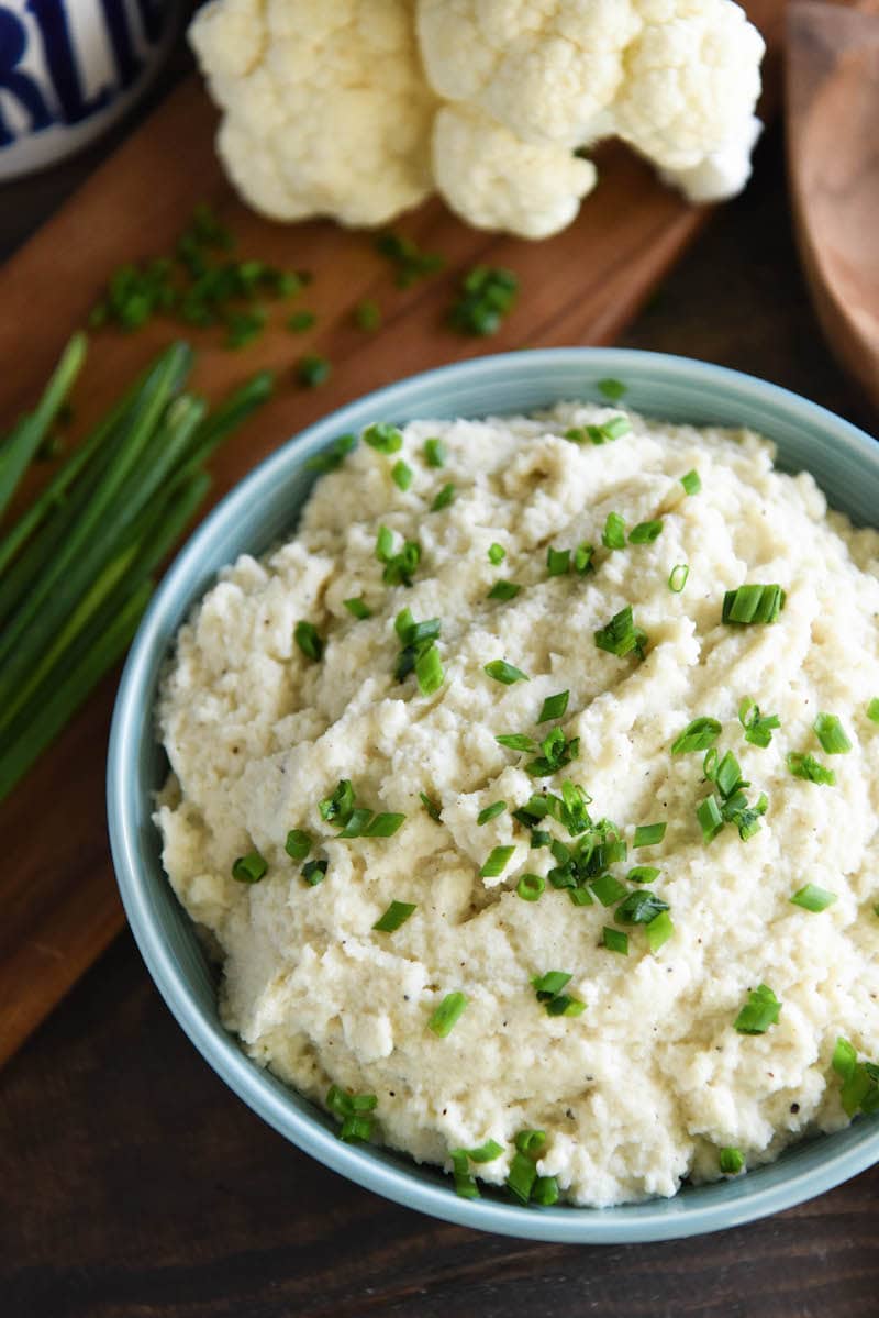 A bowl of prepared cauliflower mash next to a bunch of whole chives and cauliflower florets