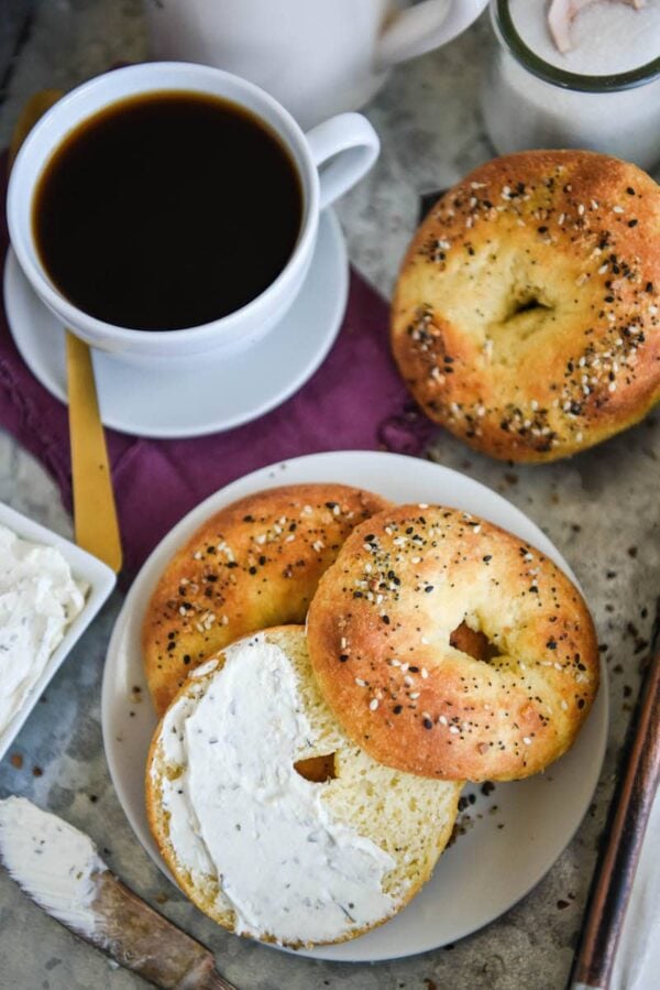 Low carb bagel sliced in half with cream cheese spread on top with a cup of coffee and a spoon.