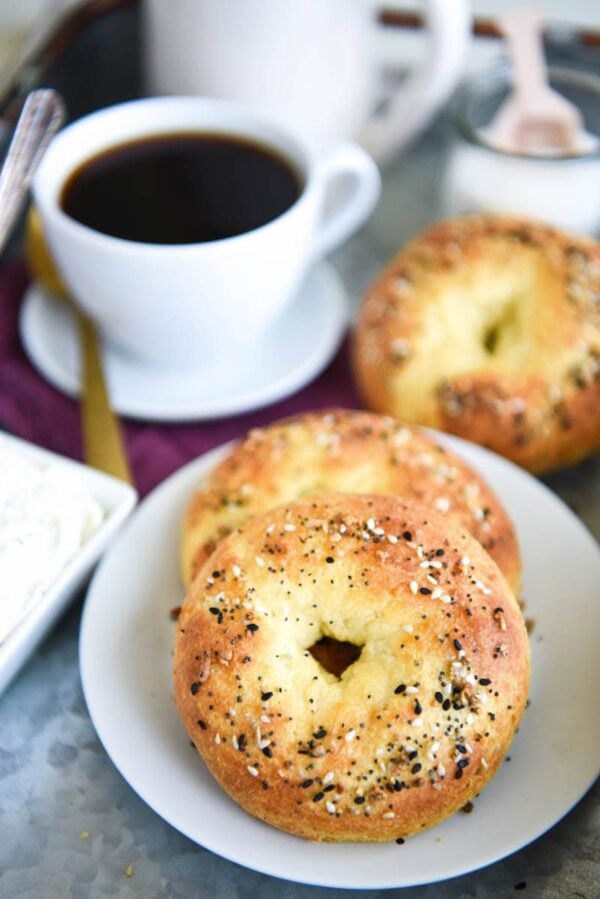 Two keto bagels on a plate with another bagel and a cup of coffee in the background.