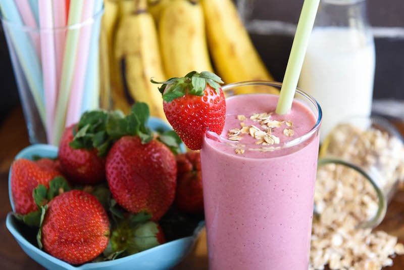 A filled smoothie glass with a bowl of strawberries, a bunch of bananas, spilled oats, and a glass of milk in the background