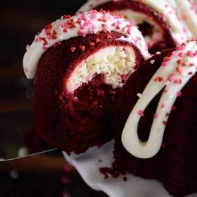 Slice of Red Velvet Cream Cheese Bundt Cake topped with red, pink, and white sprinkles being pulled from whole cake.