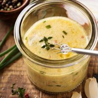 Steak sauce in a clear jar with a small whisk on a wooden board with garlic cloves