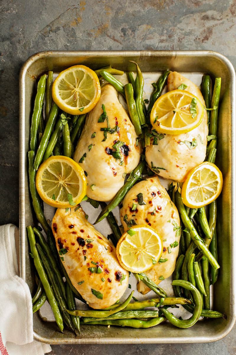 Overhead view of chicken breasts, green beans and lemons on a sheet pan.