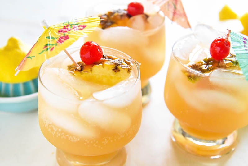 Glasses of sparkling pineapple punch garnished with cherries, pineapple slices, and a cocktail umbrella.