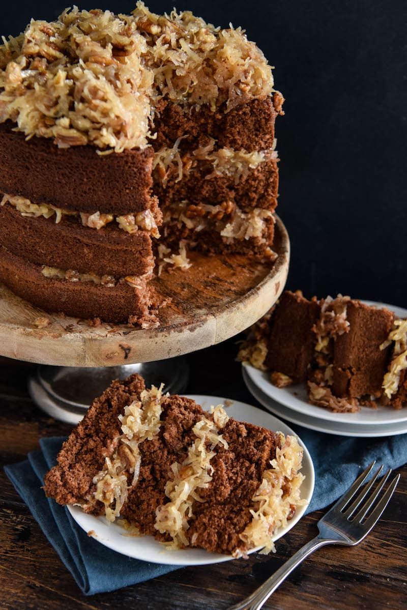 Two Plates Holding Slices of Chocolate Layer Cake Next to the Rest of the Cake on a Stand