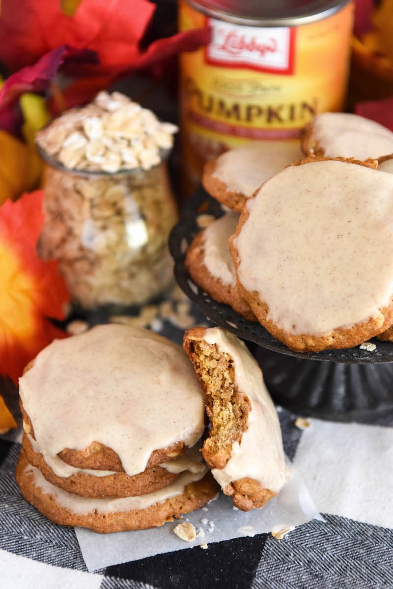 Iced pumpkin cookies in a stack with a bite missing from one, surrounded by the cookie ingredients: oats and a can of pumpkin puree