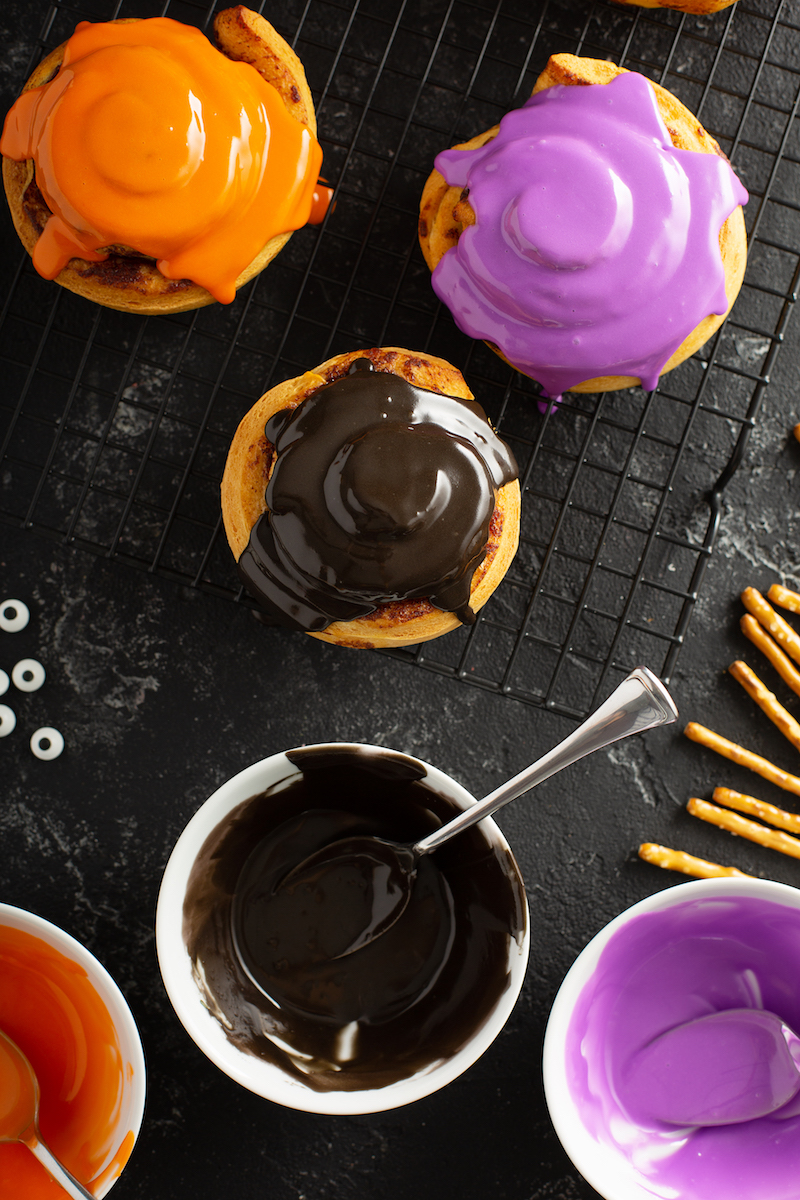 Three Iced Cinnamon Rolls For a Halloween Breakfast Beside Pretzel Sticks and Candy Eyeballs