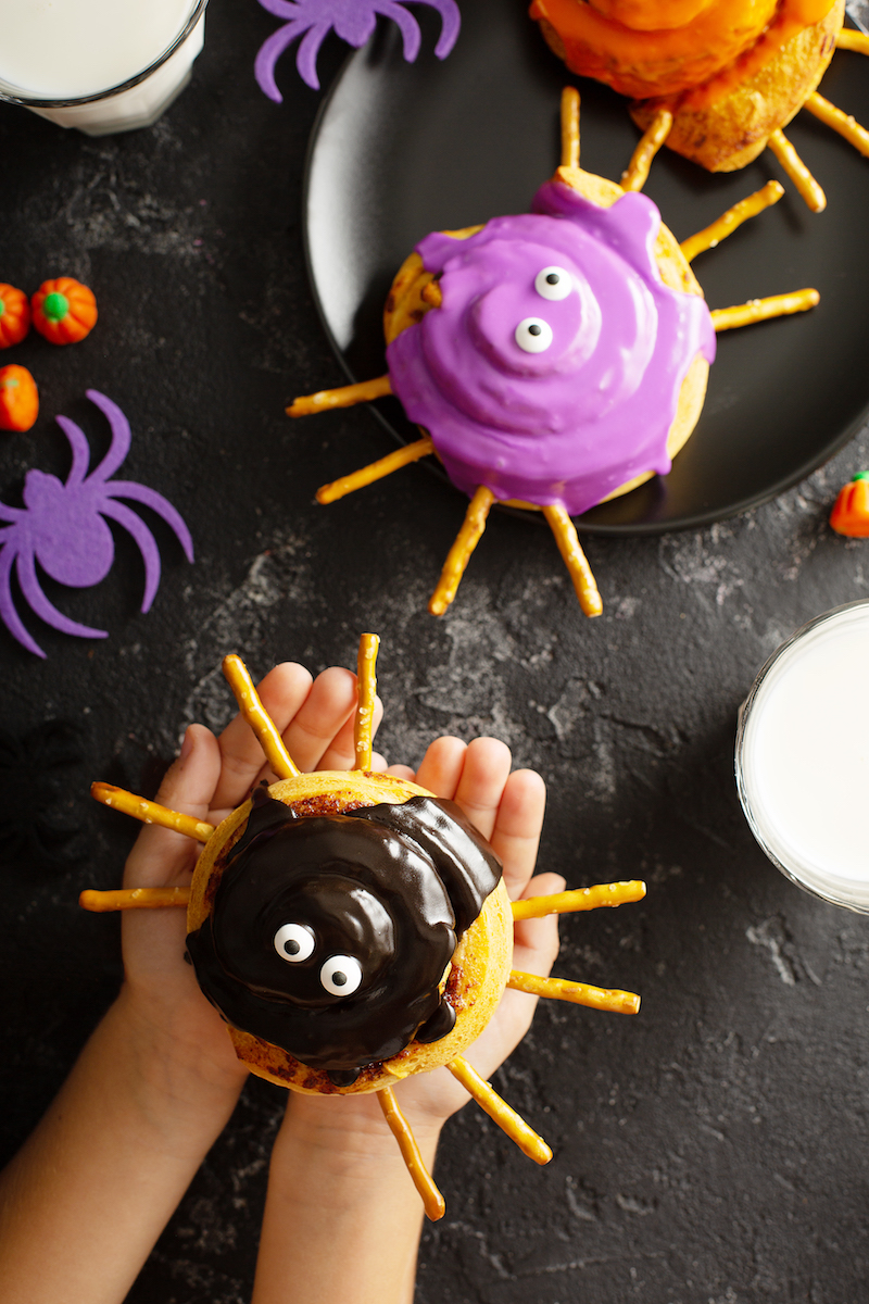 A Child's Hands Holding a Spider Cinnamon Roll with Two More Cinnamon Rolls on a Black Plate