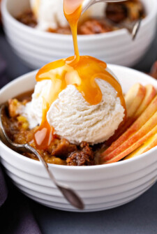 Caramel Apple Cobbler in a bowl with fresh apples on the side, vanilla ice cream on top and a caramel drizzle.