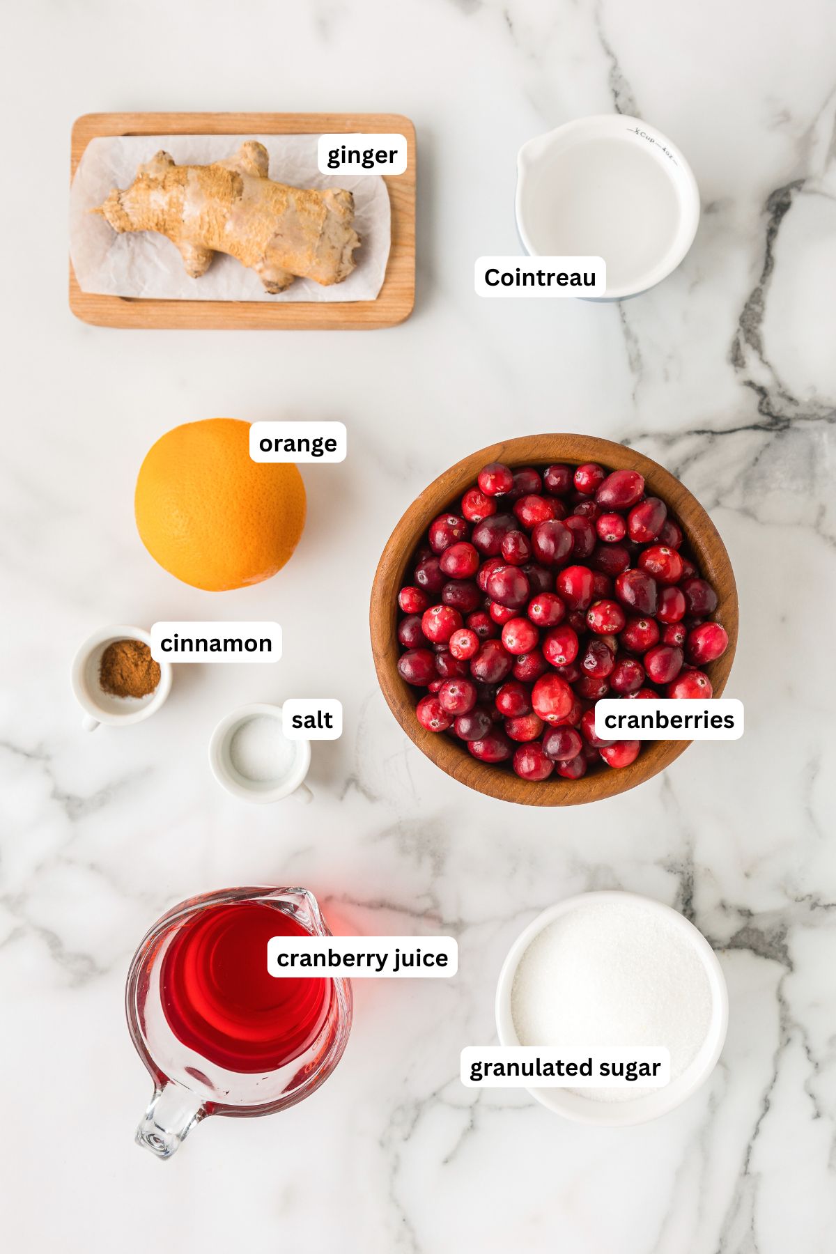 The ingredients for cranberry sauce recipe with orange in order from top to bottom: ginger, cointreau, orange, cinnamon, salt, cranberries, cranberry juice, granulated sugar. 