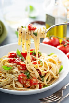 A fork with creamy fettuccine on it over a bowl of more pasta and tomatoes