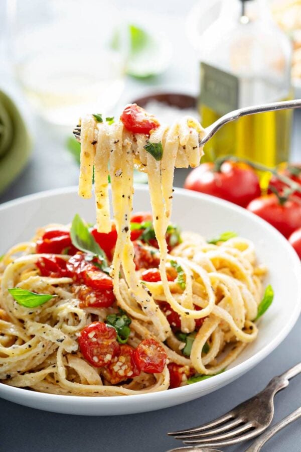 A fork with creamy fettuccine on it over a bowl of more pasta and tomatoes