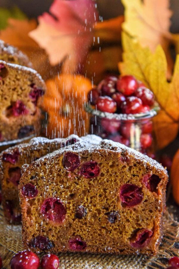 Two slices of pumpkin cranberry buttermilk bread with fresh cranberries being dusted with powdered sugar.