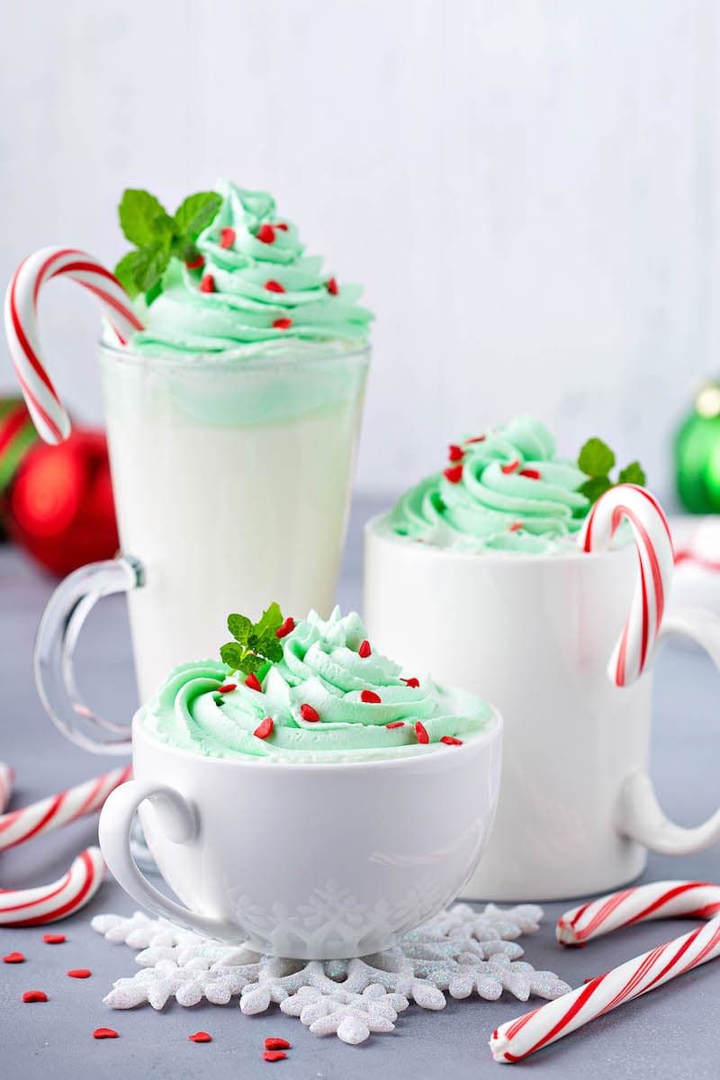 Three Different-Sized Mugs of Peppermint White Hot Chocolate with Green Whipped Cream and Candy Canes Placed Inside