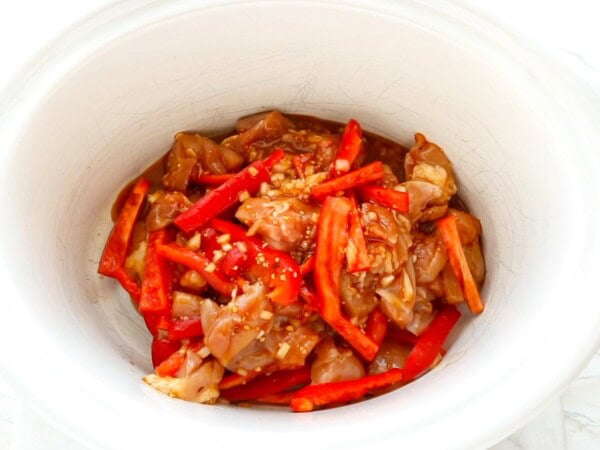 Raw chicken and red peppers are coated in sesame sauce in a crockpot. 