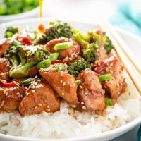 Close up shot of sesame chicken with broccoli served over white rice topped with green onions and sesame seeds.