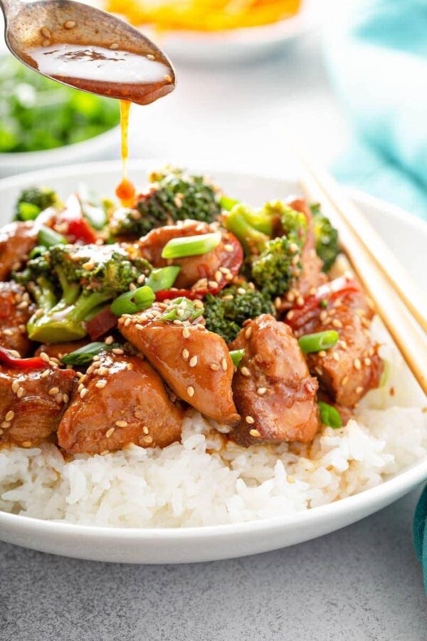 Close up shot of sesame chicken with broccoli served over white rice topped with green onions and sesame seeds. 