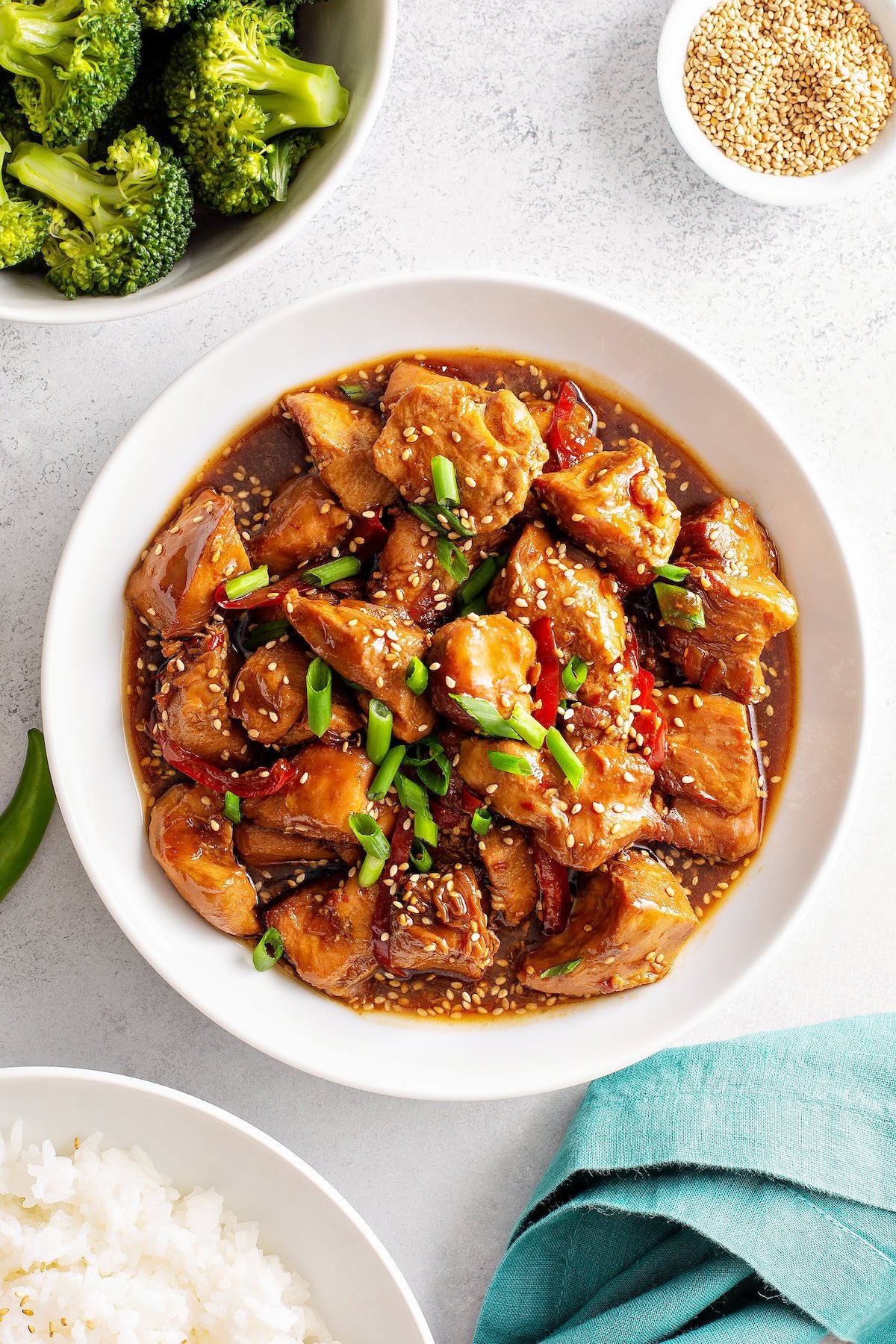 Saucy sesame chicken is presented on a bowl, garnished with chopped green onions and sesame seeds. 