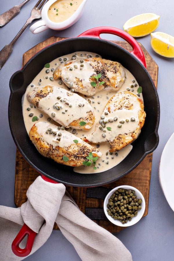 Chicken breasts covered in lemon caper cream sauce in a large cast iron skillet.