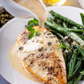 Sautéed chicken breast and green beans with a creamy lemon caper sauce being poured on top.