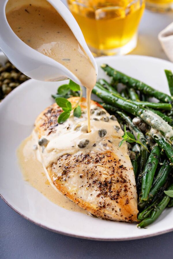 Sautéed chicken breast and green beans with a creamy lemon caper sauce being poured on top. 