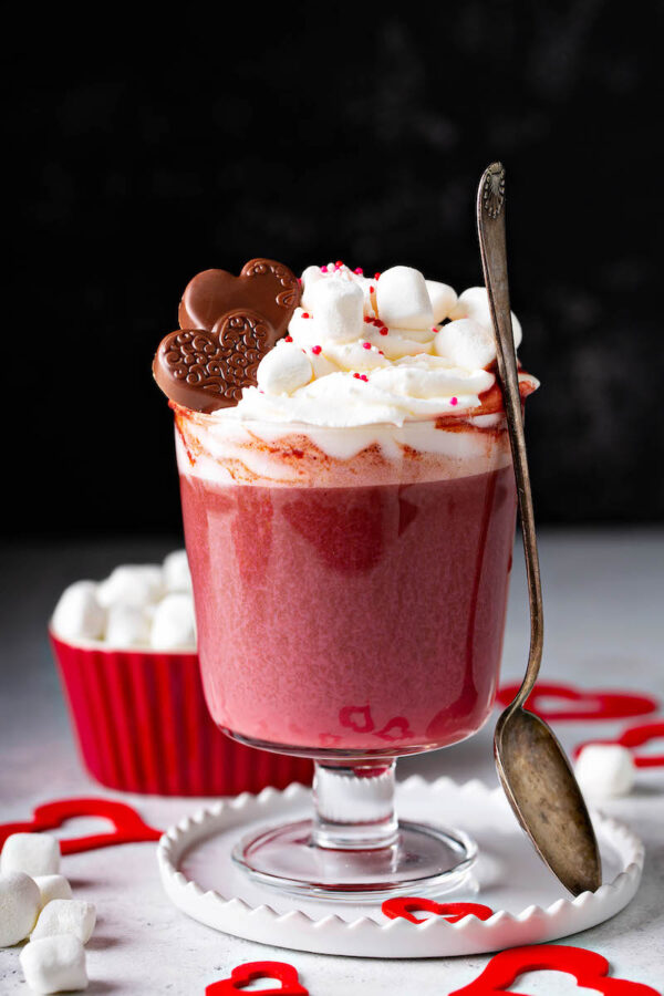 Red Velvet Hot Chocolate in a glass mug with marshmallow whip cream on top and a spoon to stir. 