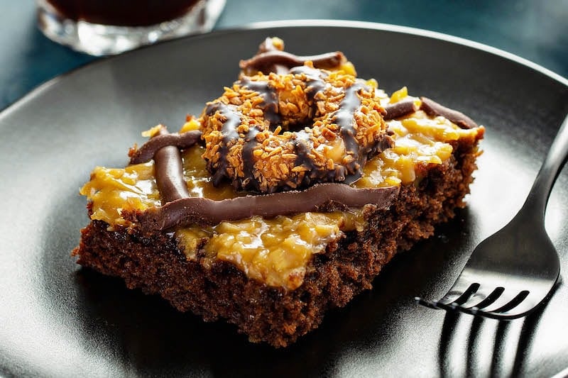 Piece of Samoa Cookies Sheet Cake topped with Samoa Girl Scout Cookie set in a black plate