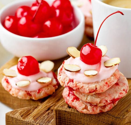 Cherry Almond Sugar Cookies stacked together with maraschino cherries on a cutting board.