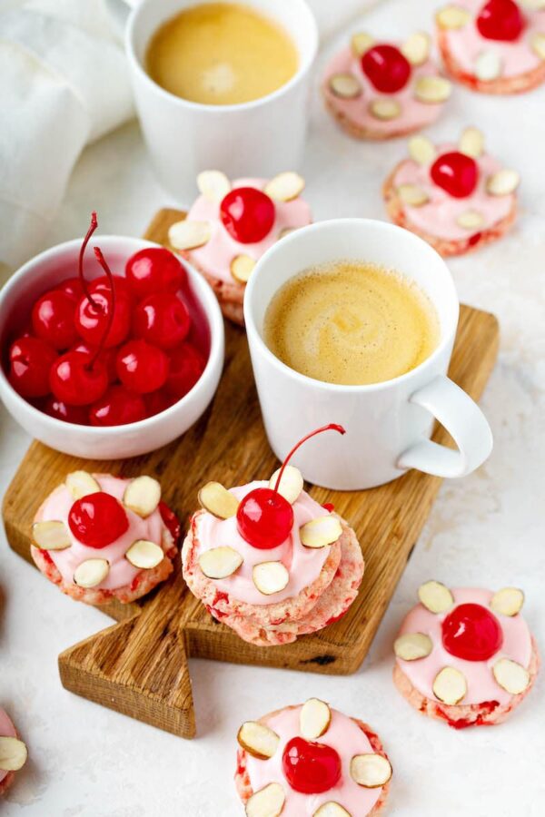 Maraschino Cherries on top of Cherry Almond Cookies with pink frosting, a cup of coffee and maraschino cherries in a white bowl.