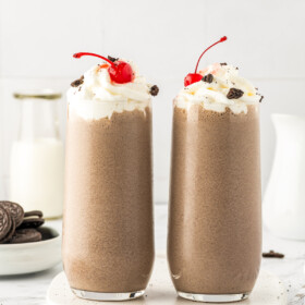 Two milkshakes, side by side, topped with whipped cream and cherries.