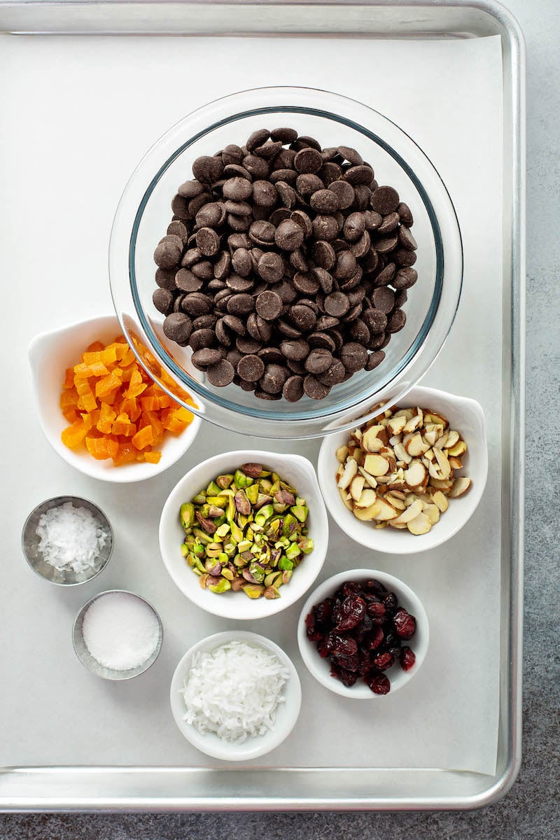 A Lined Baking Sheet Holding Bowls of Chocolate Chips, Coconut Flakes and the Remaining Bark Ingredients