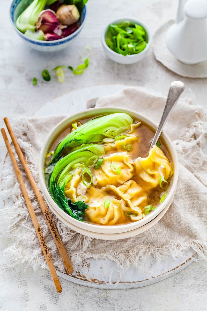 Wonton soup in a white bowl with chopsticks and a spoon.