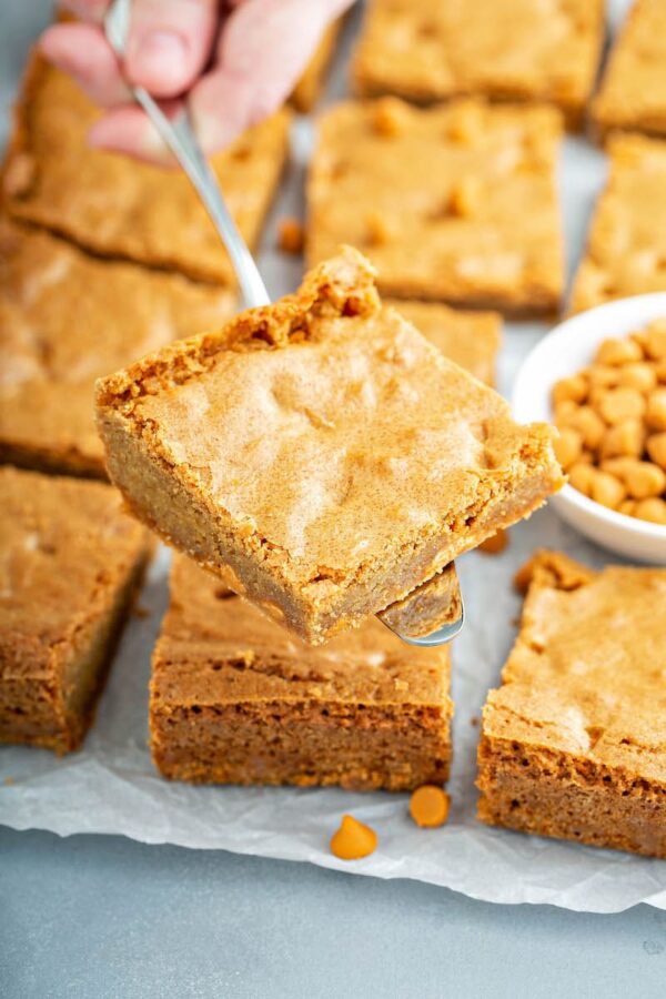 Butterscotch Blondies on parchment paper and one is being scooped up with a spatula. 