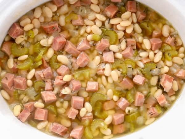 A white crockpot is filled with beans, ham, and celery.