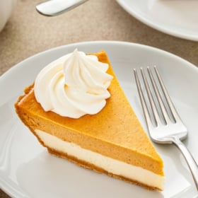 Whipped cream is piped on top of a slice of pumpkin pie cheesecake.