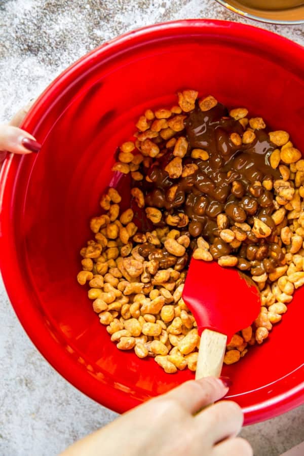 Corn cereal is being mixed with chocolate and peanut butter in a large red bowl.