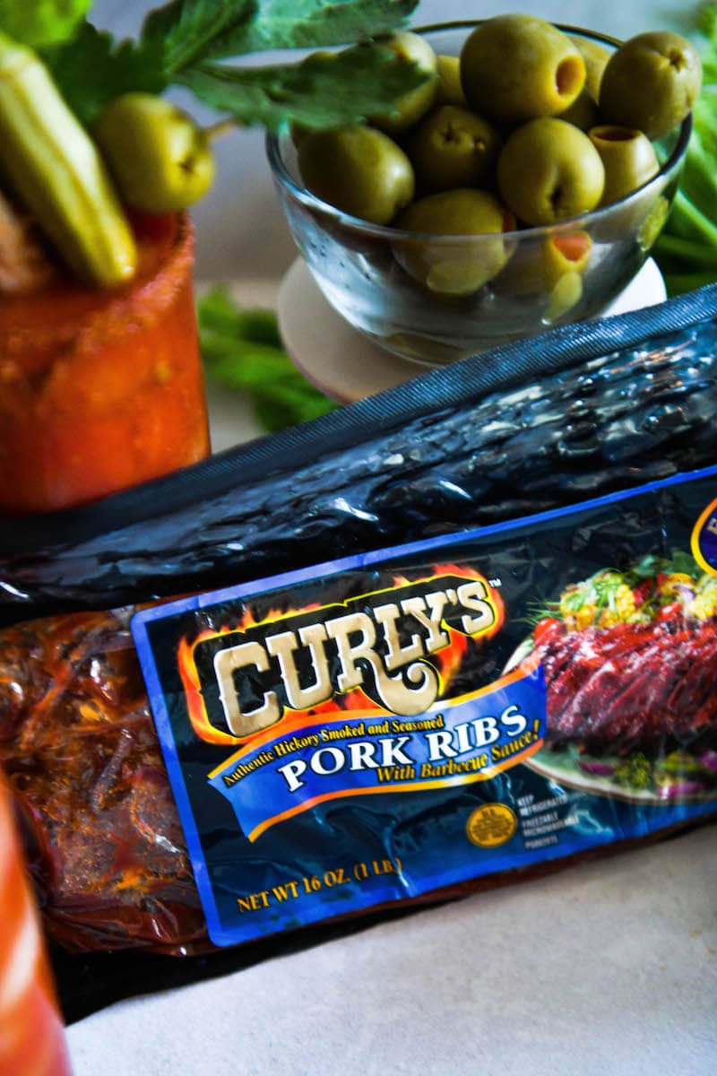 A closeup of Curly’s Baby Back Ribs, the garnish of choice for this recipe, alongside a bowl of olives and a glass of tomato mix.